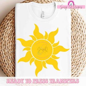 May include: A white t-shirt with a yellow sun design. The sun has a circle in the center with the text "Suzy Q Designs" inside. The text "Ready to Press Transfers" is at the bottom of the image.