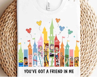 May include: A white t-shirt with a colorful graphic of the Toy Story characters in front of a rainbow-colored castle silhouette. The text "You've got a friend in me" is printed below the graphic. The shirt is ready to press.