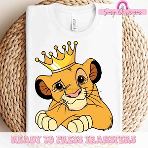 May include: A white t-shirt with a cartoon image of a lion cub wearing a gold crown. The lion cub is looking to the side with a serious expression. The text "READY TO PRESS TRANSFERS" is printed at the bottom of the shirt.