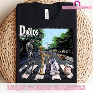 Puede incluir: Una camiseta negra con un gr&aacute;fico de personajes de Star Wars, R2-D2, C-3PO y BB-8, caminando por un paso de cebra, similar a la portada del &aacute;lbum Abbey Road de los Beatles. El texto "The Droids" y "Imperial Road" est&aacute; por encima de los personajes.