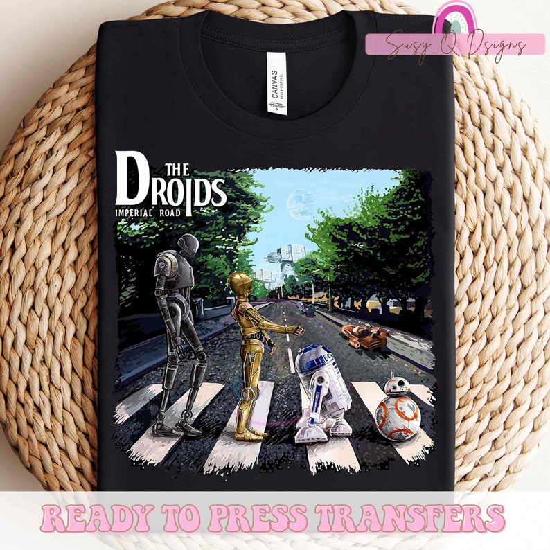 Puede incluir: Una camiseta negra con un gr&aacute;fico de personajes de Star Wars, R2-D2, C-3PO y BB-8, caminando por un paso de cebra, similar a la portada del &aacute;lbum Abbey Road de los Beatles. El texto "The Droids" y "Imperial Road" est&aacute; por encima de los personajes.