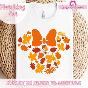 May include: White t-shirt featuring a fall-themed design. The design includes an orange bow, leaves, acorns, and hearts arranged in a Mickey Mouse head shape. The text "Matching Set" and "READY TO PRESS TRANSFERS" are also visible.