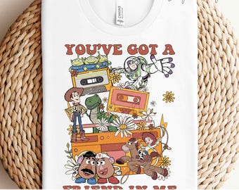 May include: White t-shirt with a colourful graphic design featuring characters from the Toy Story film. The design includes the text "YOU'VE GOT A FRIEND IN ME" and "READY TO PRESS TRANSFERS". The design includes Woody, Buzz Lightyear, and other characters.