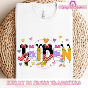May include: A white t-shirt with a design featuring the name "JAIDEN" in colorful letters with Mickey Mouse and Minnie Mouse characters. The design also includes other Disney characters like Pluto and Daisy Duck. The text "READY TO PRESS TRANSFERS" is at the bottom of the shirt.
