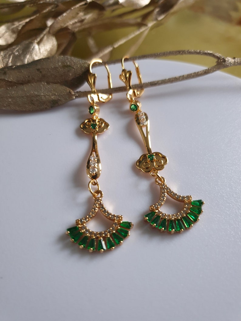 Op de afbeelding: Gouden oorhangers met groene en witte stenen. De oorbellen hebben een waaiervormig ontwerp met een wolkvormige hanger boven de waaier.