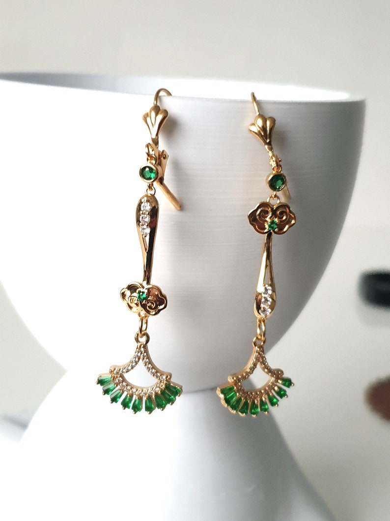 Op de afbeelding: Gouden oorhangers met groene en heldere kristallen accenten. De oorbellen hebben een wolkontwerp en een waaiervormige onderkant.