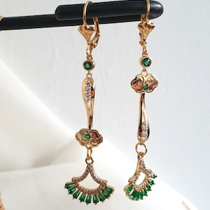 Op de afbeelding: Een paar gouden oorhangers met groene en heldere kristallen accenten. De oorbellen hebben een wolkvormig ontwerp met een waaiervormige hanger.
