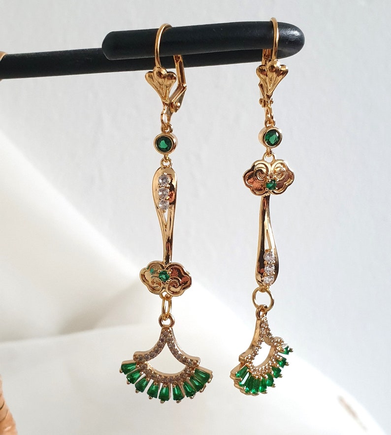 Op de afbeelding: Een paar gouden oorhangers met groene en heldere kristallen accenten. De oorbellen hebben een wolkvormig ontwerp met een waaiervormige hanger.