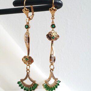 Op de afbeelding: Een paar gouden hangende oorbellen met groene en heldere kristallen accenten. De oorbellen hebben een wolkvormig ontwerp met een waaiervormige hanger.
