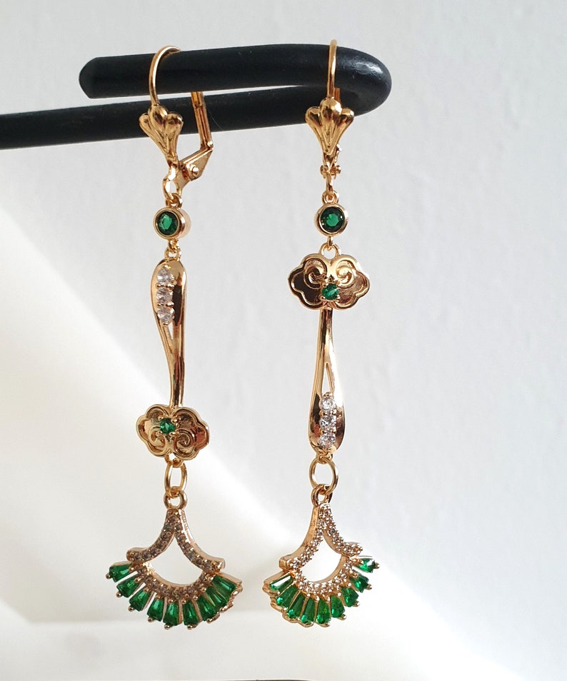 Op de afbeelding: Een paar gouden hangende oorbellen met groene en heldere kristallen accenten. De oorbellen hebben een wolkvormig ontwerp met een waaiervormige hanger.