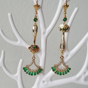 Op de afbeelding: Gouden oorhangers met groene en heldere kristallen accenten. De oorbellen hebben een wolkontwerp en een waaiervormige onderkant.