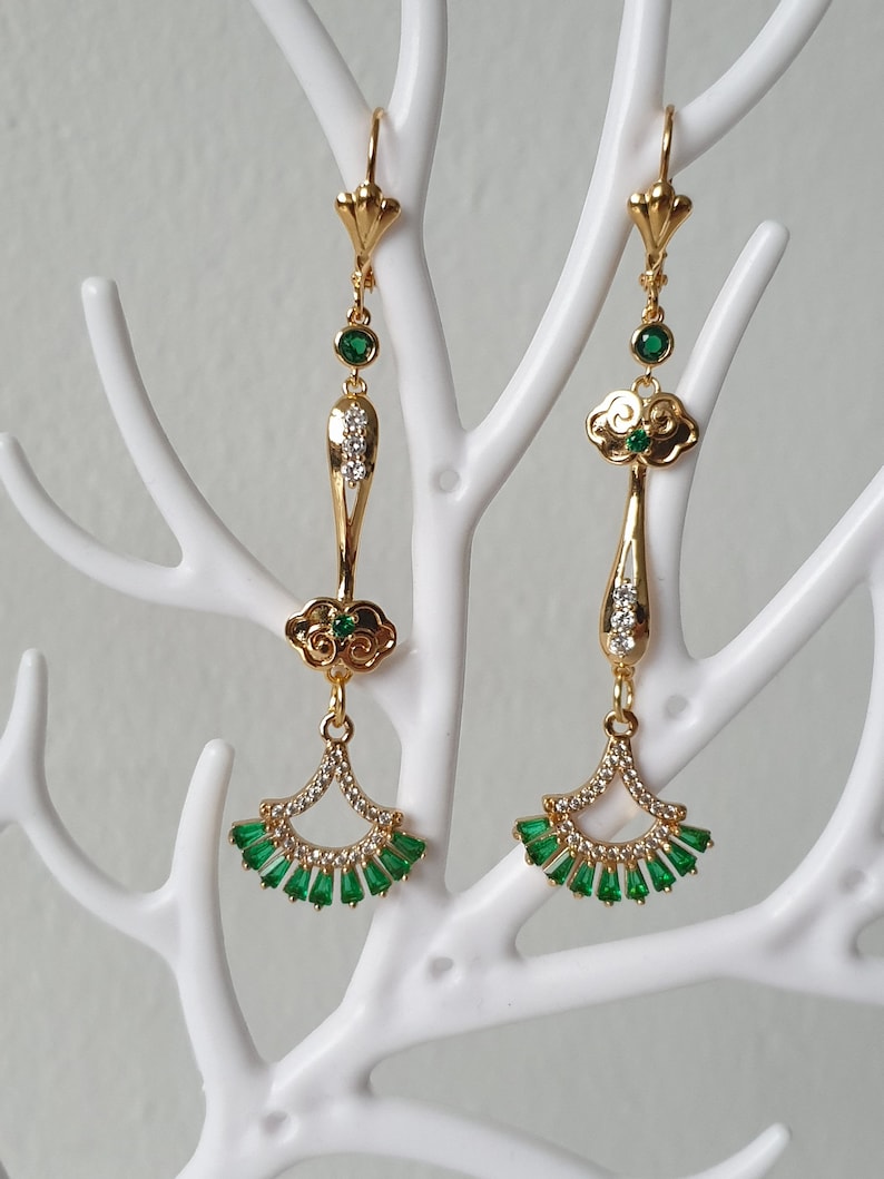 Op de afbeelding: Gouden oorhangers met groene en heldere kristallen accenten. De oorbellen hebben een wolkontwerp en een waaiervormige onderkant.
