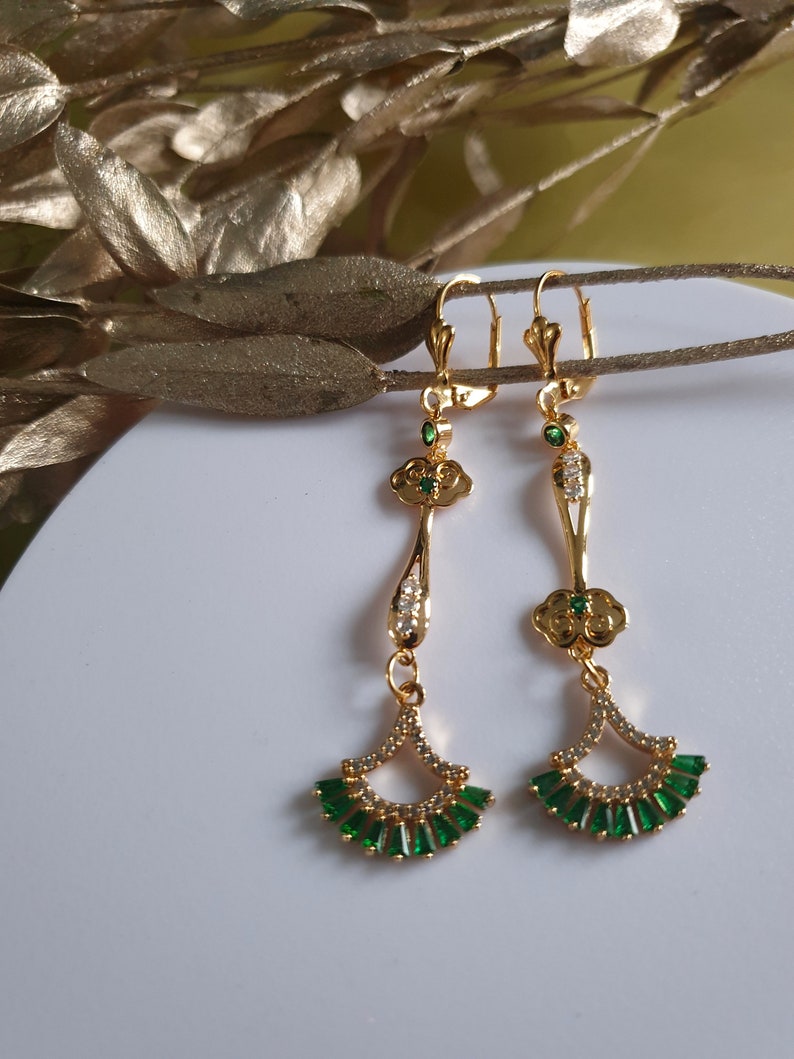 Op de afbeelding: Gouden oorhangers met groene en witte stenen. De oorbellen hebben een waaiervormig ontwerp met een klein bloemetjesdetail.