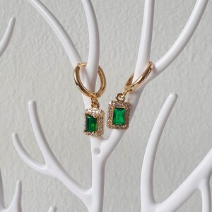 May include: Gold earrings with a green emerald-shaped stone set in a clear crystal halo.