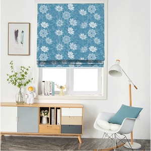 May include: A blue roman blind with a white floral pattern hangs in a window. The blind is raised, revealing a white window frame and a view of a white wall. A wooden floor is visible below the window.