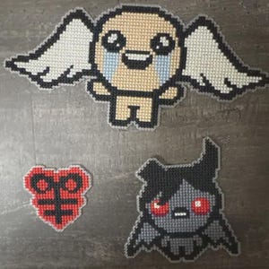 Peut inclure: Trois motifs en pixel art : un ange avec des ailes, un cœur rouge avec une croix noire et une créature en forme de chauve-souris avec des yeux rouges. L'ange a un corps beige, un contour noir et des ailes blanches.
