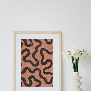 May include: A framed print featuring an abstract pattern in black and orange. The print is in a light wood frame and is displayed on a white surface. A vase with white flowers is positioned to the right of the print.