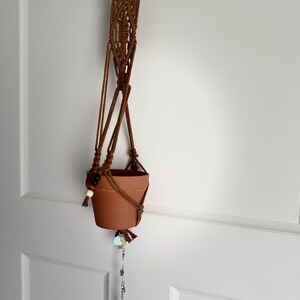 May include: A brown macrame plant hanger with a terracotta pot. The hanger features a wooden ring at the top and a decorative beaded accent. A crystal and red stone dangle from the bottom, adding a touch of sparkle. The pot is approximately 4 inches in diameter.