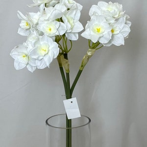 May include: A bouquet of white artificial daffodils with yellow centers in a clear glass vase. The flowers have a realistic appearance and are arranged in a cluster.