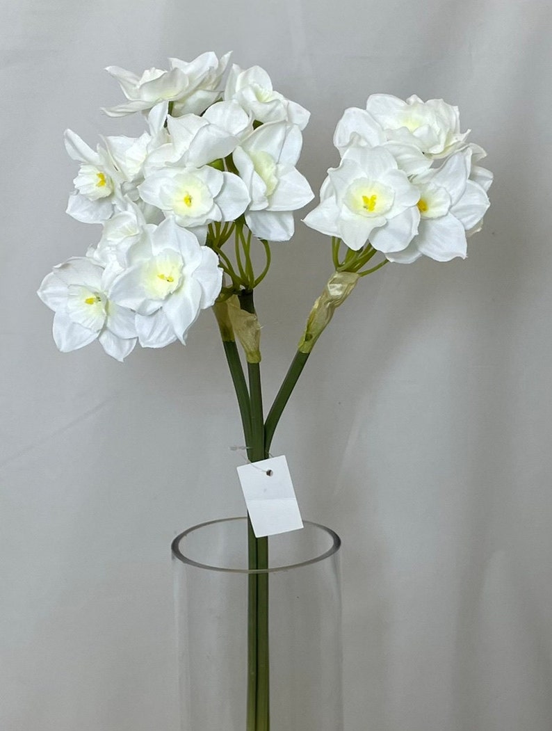 May include: A bouquet of white artificial daffodils with yellow centers in a clear glass vase. The flowers have a realistic appearance and are arranged in a cluster.