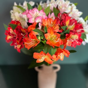 May include: A bouquet of orange, pink, and white alstroemeria flowers in a white ceramic vase. The flowers are arranged in a loose, natural style.
