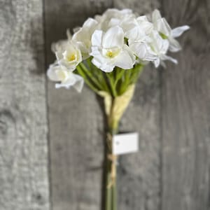 May include: A bouquet of white artificial daffodils with green stems and leaves. The flowers are arranged in a cluster and have a realistic appearance.