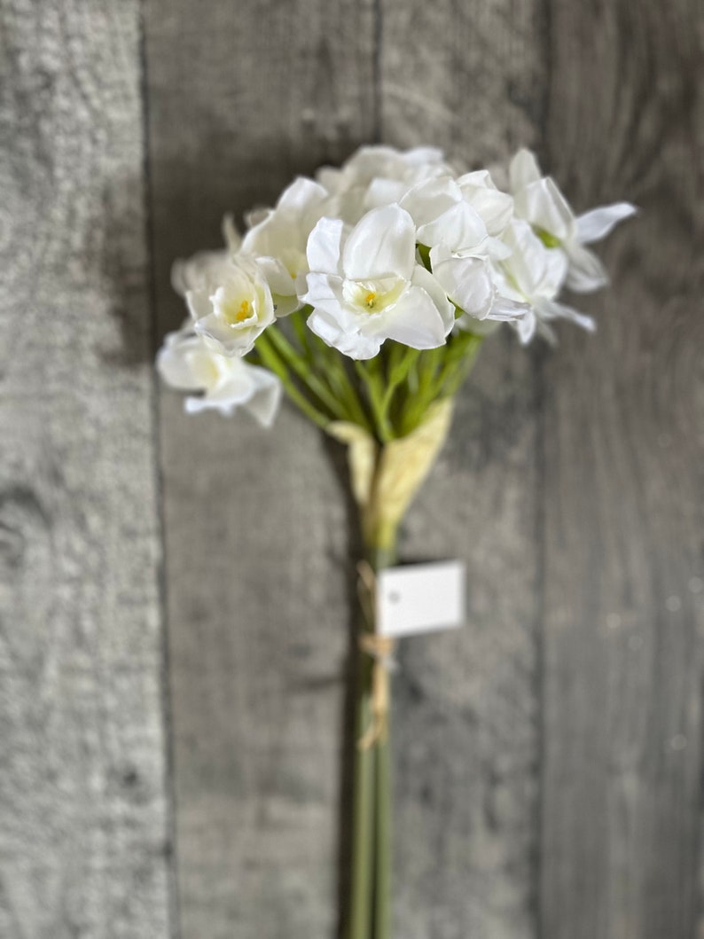 May include: A bouquet of white artificial daffodils with green stems and leaves. The flowers are arranged in a cluster and have a realistic appearance.