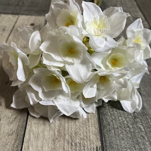 May include: A bouquet of white silk daffodils with yellow centers. The flowers are arranged in a cluster and have a realistic appearance.