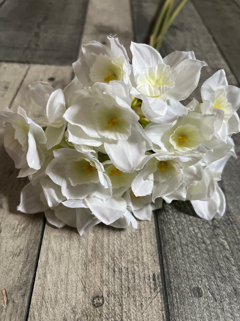 May include: A bouquet of white silk daffodils with yellow centers. The flowers are arranged in a cluster and have a realistic appearance.