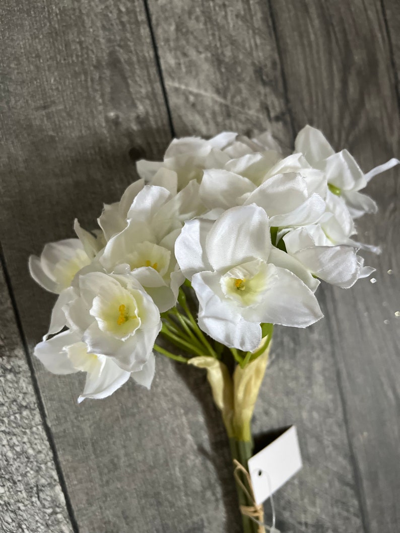 May include: A bouquet of white artificial daffodils with green stems. The flowers are arranged in a cluster and have a realistic appearance.