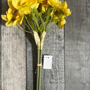 May include: A bouquet of yellow daffodils with green stems tied together with a brown ribbon. The flowers are arranged in a cluster and have a natural, rustic look.