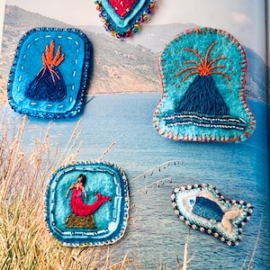 May include: Five embroidered patches featuring a red heart with gold rays, two volcanoes with orange lava, a mermaid with a red tail, and a blue fish. Each patch is crafted with colorful thread, beads, and felt, showcasing intricate needlework details.