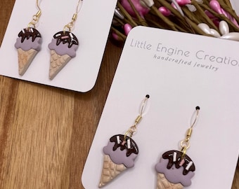 Puede incluir: Pendientes colgantes de cono de helado morado y marrón con ganchos dorados. Los pendientes están en tarjetas blancas con el texto "Little Engine Creations handcrafted jewelry".