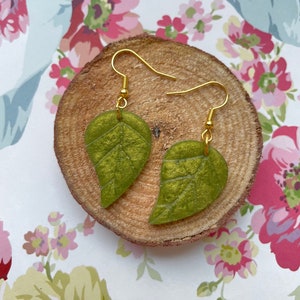 May include: A pair of green leaf-shaped earrings with gold hooks. The earrings are displayed on a wooden slice.