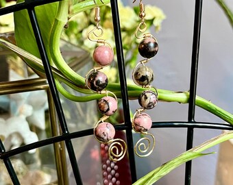 May include: A pair of gold wire wrapped earrings with pink and black gemstone beads. The earrings are hanging from a black wire rack.