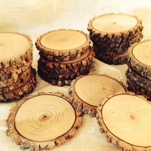 May include: A collection of natural wood slices, each with a unique bark pattern and a smooth, light brown surface. The slices are stacked in groups of varying heights, creating a rustic and organic aesthetic.
