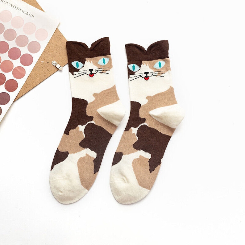 May include: A pair of cat-themed socks. The socks feature a camouflage pattern in shades of brown, tan, and white. Each sock has a cat face design at the top, with blue eyes, a small red mouth, and brown ears.