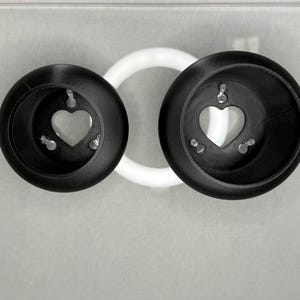 May include: Two black, circular objects with heart-shaped cutouts and small silver accents. A white, circular object is positioned between them. The objects appear to be decorative or functional, possibly for home decor or organization.