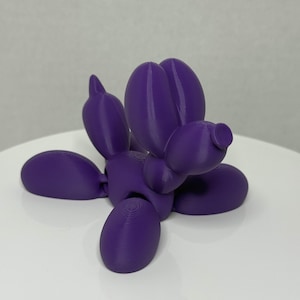 Puede incluir: Una escultura de animal de globo morado, que se asemeja a un perro, se exhibe sobre una superficie blanca. La escultura está hecha de un material sólido y presenta formas redondeadas, con cabeza, cuerpo, patas y cola. El color es un morado vibrante y sólido.