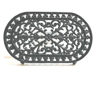 May include: An oval-shaped black cast iron trivet featuring an intricate openwork scroll and floral pattern. The design includes a decorative border with repeating leaf-like motifs and a central symmetrical arrangement of curved metal vines.