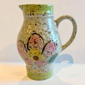 Puede incluir: Jarra de cerámica con diseño floral. La jarra tiene un cuerpo beige moteado con un diseño de flores rosas y amarillas. El borde y la base son de color verde claro. Tiene un asa curva y un pico para verter.