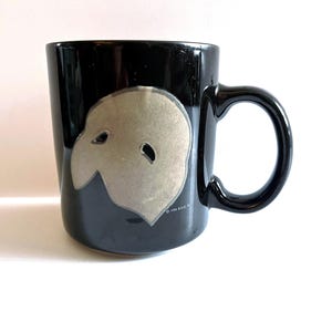 May include: Black ceramic mug featuring a gray mask design. The mug has a rounded handle and a glossy finish. The mask design is a light gray color and is centered on the mug. The mug is on a white surface.