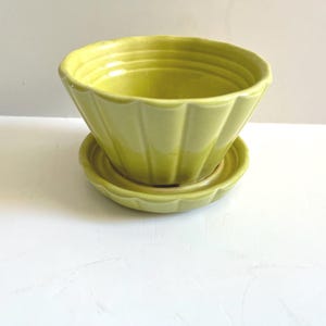May include: A chartreuse ceramic planter with a matching saucer. The planter has a scalloped rim and vertical ribbed detailing. The saucer is round with a matching scalloped edge. The set is on a white surface.