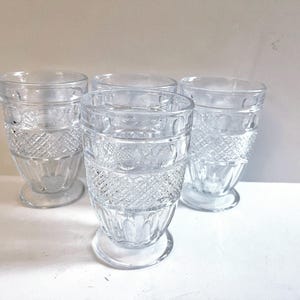 May include: Four clear, vintage-style drinking glasses. Each glass features a decorative diamond pattern around the base and a smooth rim. The glasses are made of clear glass and are displayed on a white surface.