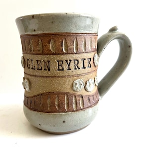 May include: Handmade ceramic mug with a gray, brown, and tan color scheme. The mug features the text "GLEN EYRIE" and decorative embossed details. The handle is curved and the mug has a rustic, artisanal appearance.