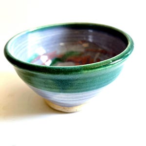 May include: A handmade ceramic bowl with a rounded shape. The bowl features a green rim, a blue-green exterior, and a light blue interior. The base is a light beige color. The bowl is suitable for serving food.