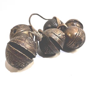 May include: A cluster of five spherical metal bells with a dark, weathered bronze finish. Each bell features an intricate embossed scrollwork pattern and a horizontal slit opening. The bells are connected by a thin, light-colored cord.