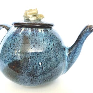 May include: A blue and brown ceramic teapot with a unique, speckled glaze. The teapot has a round body and a curved spout. The lid is topped with a small, light-colored rock.