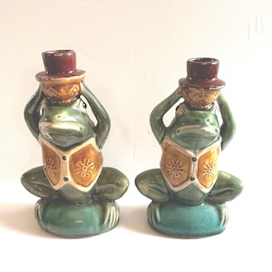 May include: Two ceramic frog figurines, each with a green body, a brown top hat, and a tan vest with a floral design. The frogs are seated on a green cushion, creating a whimsical decorative item.