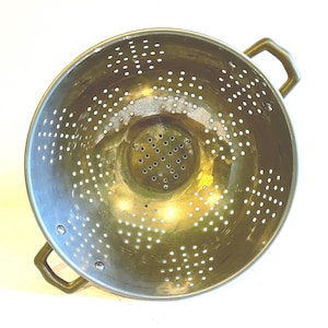 May include: A vintage brass colander with numerous small holes for draining. The colander has two handles and a rounded shape. The brass has a warm, golden hue, and the colander is set against a white background.